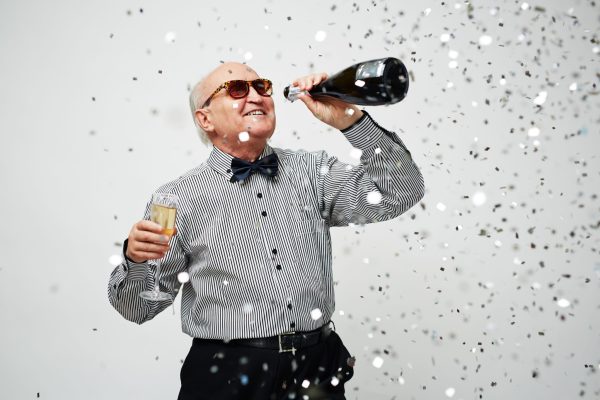 Portrait of dressed-up male senior hanging out with a bottle of champagne as confetti falling down on white background